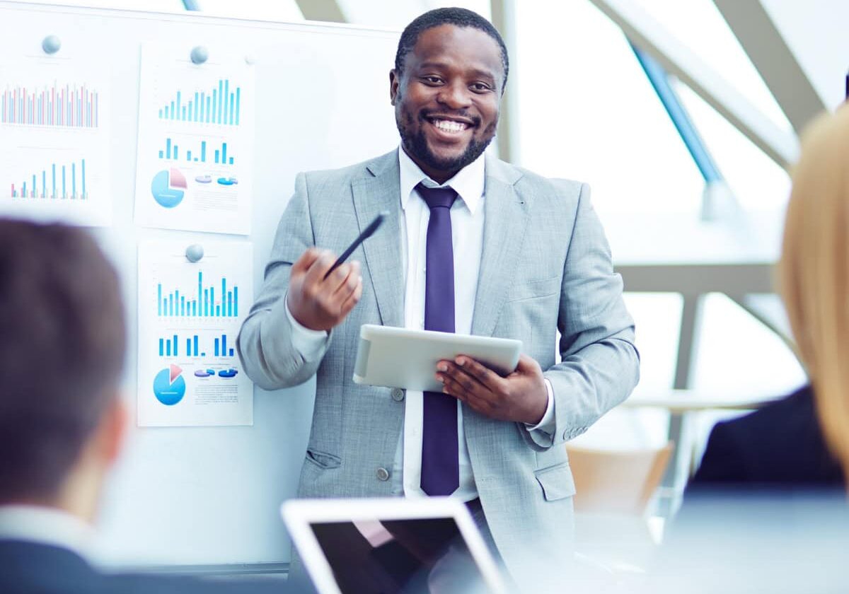 Research_Survey_Data A smiling man holding a tablet and presenting data to a group of colleagues